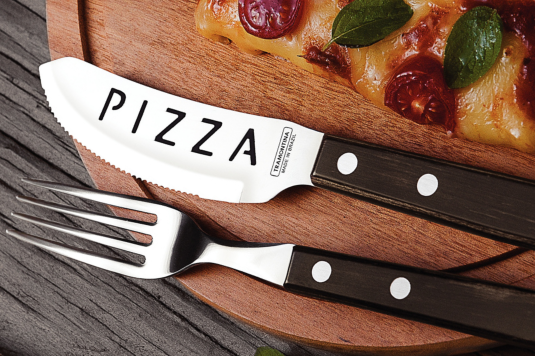 This is an image of a pizza knife and fork on the edge of a wooden plate with a pizza slice in the top right hand corner of the plate