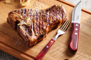 This is an image of a steak knife and fork and both of them have a very beautiful dark red wooden handle and there is a T-bone steak off to the left